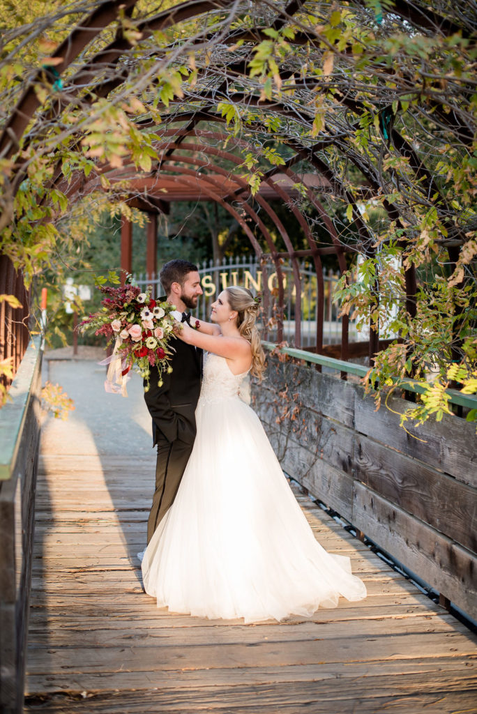 Blonde blue eyed bride and groom standing in each others arms on a plant covered bridge at Casa Bella in Sunol CA