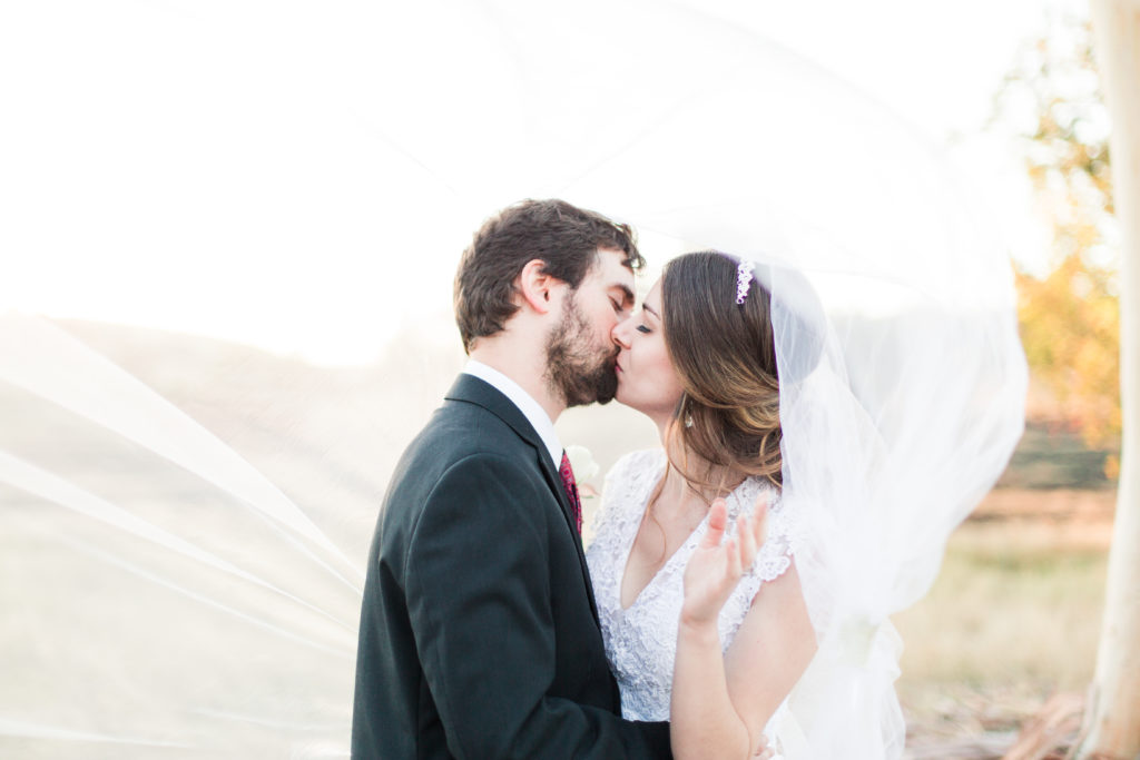 Brunette bride with classic bridal makeup kissing her groom under a veil blowing in the wind at San Jose, CA